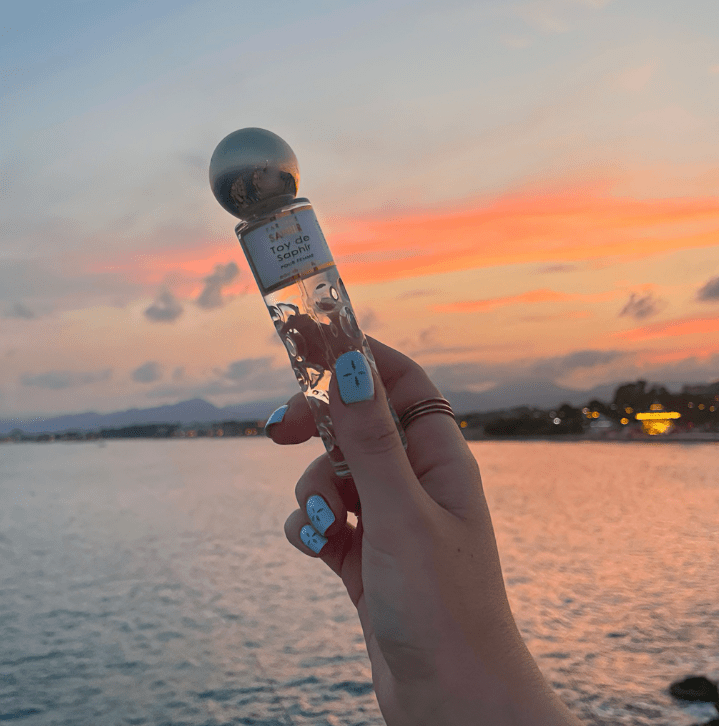 Frasco de perfume sostenido por una mano con manchas azules, frente a un atardecer colorido sobre el mar.